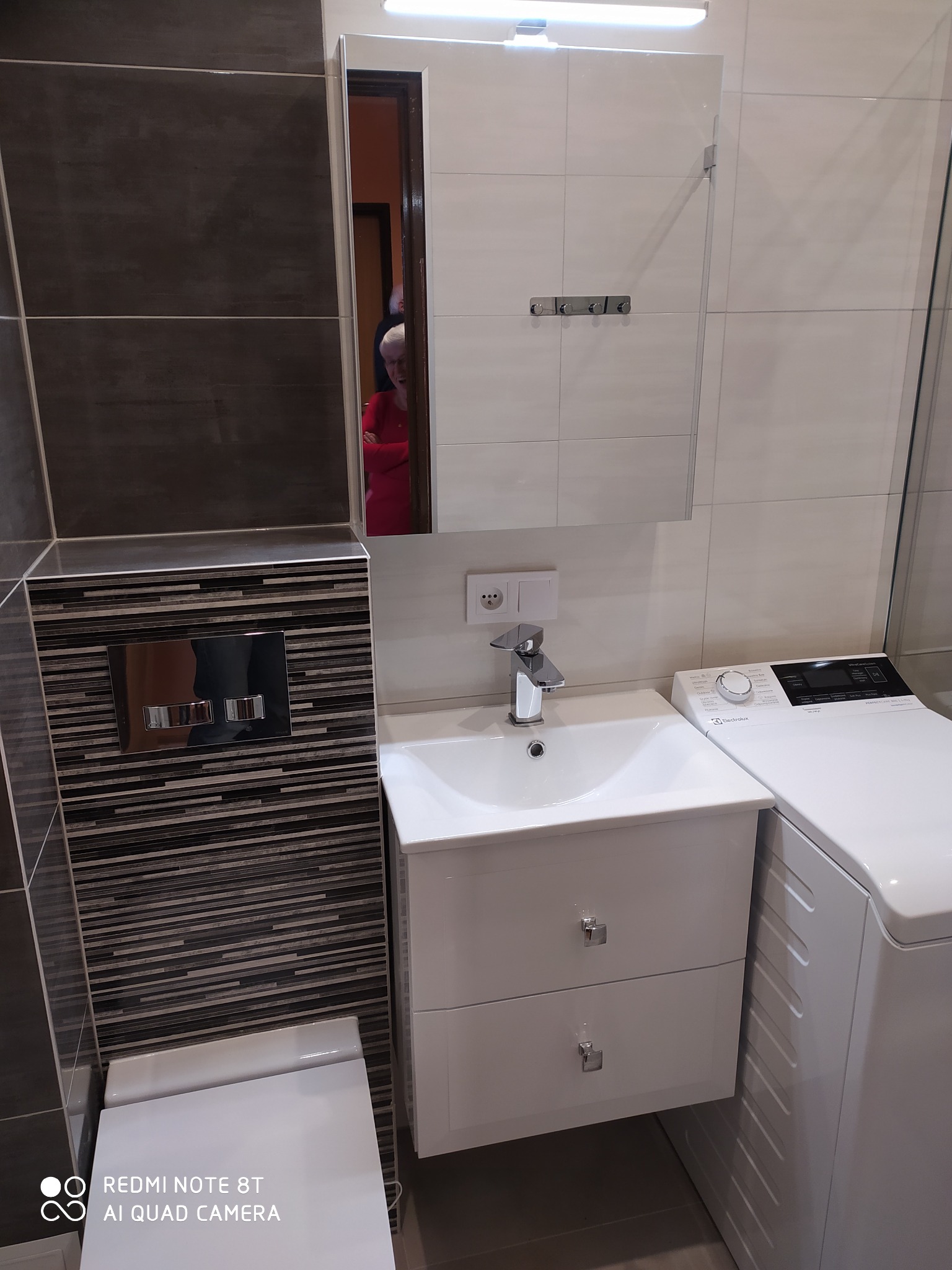 Nowoczesna łazienka po remoncie z białą umywalką z szafką, pralką Electrolux i toaletą podwieszaną z czarno-szarymi płytkami dekoracyjnymi.