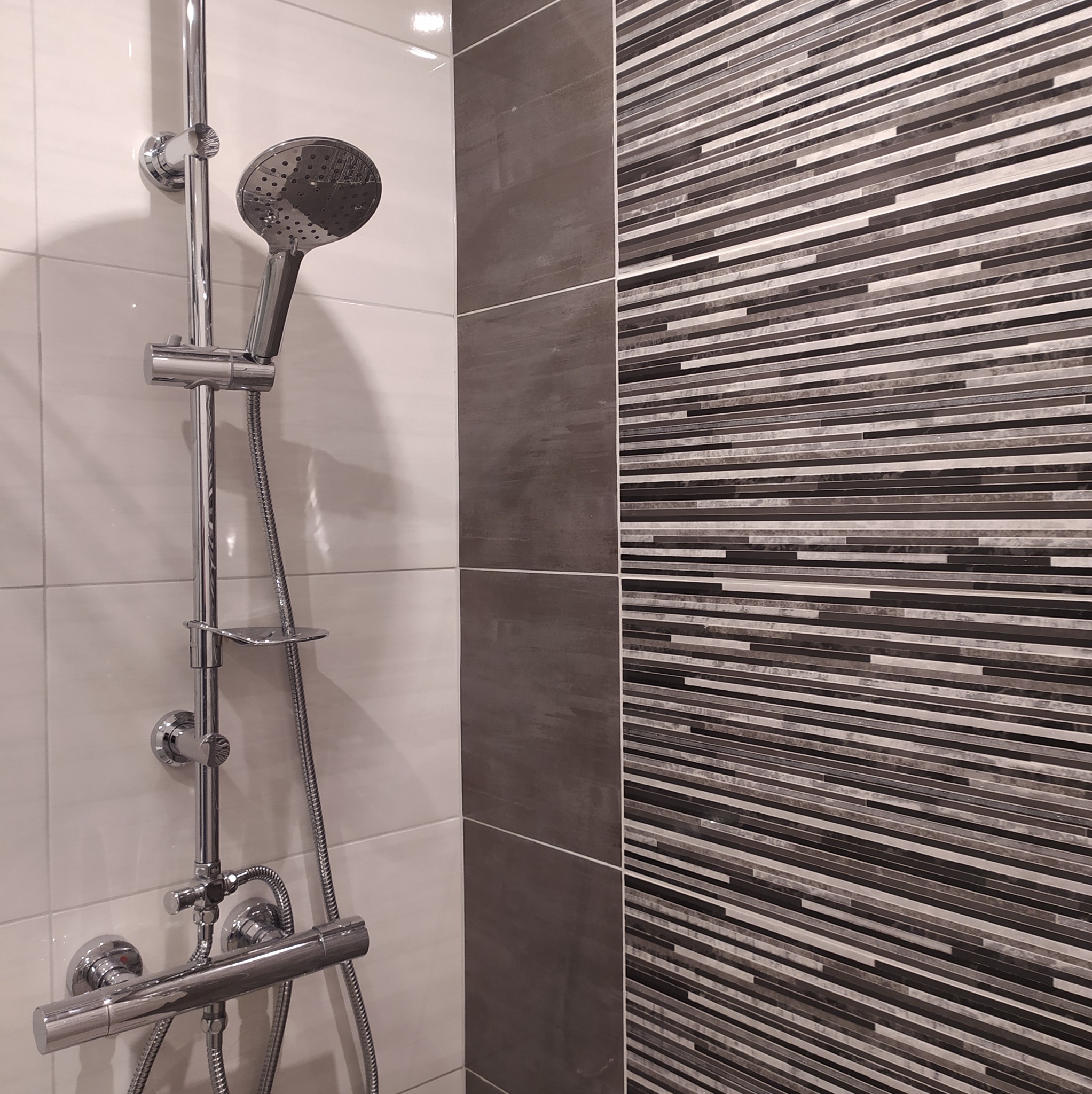 Bateria prysznicowa z deszczownicą na tle ściany wyłożonej biało-szaro-brązowymi płytkami w paski.