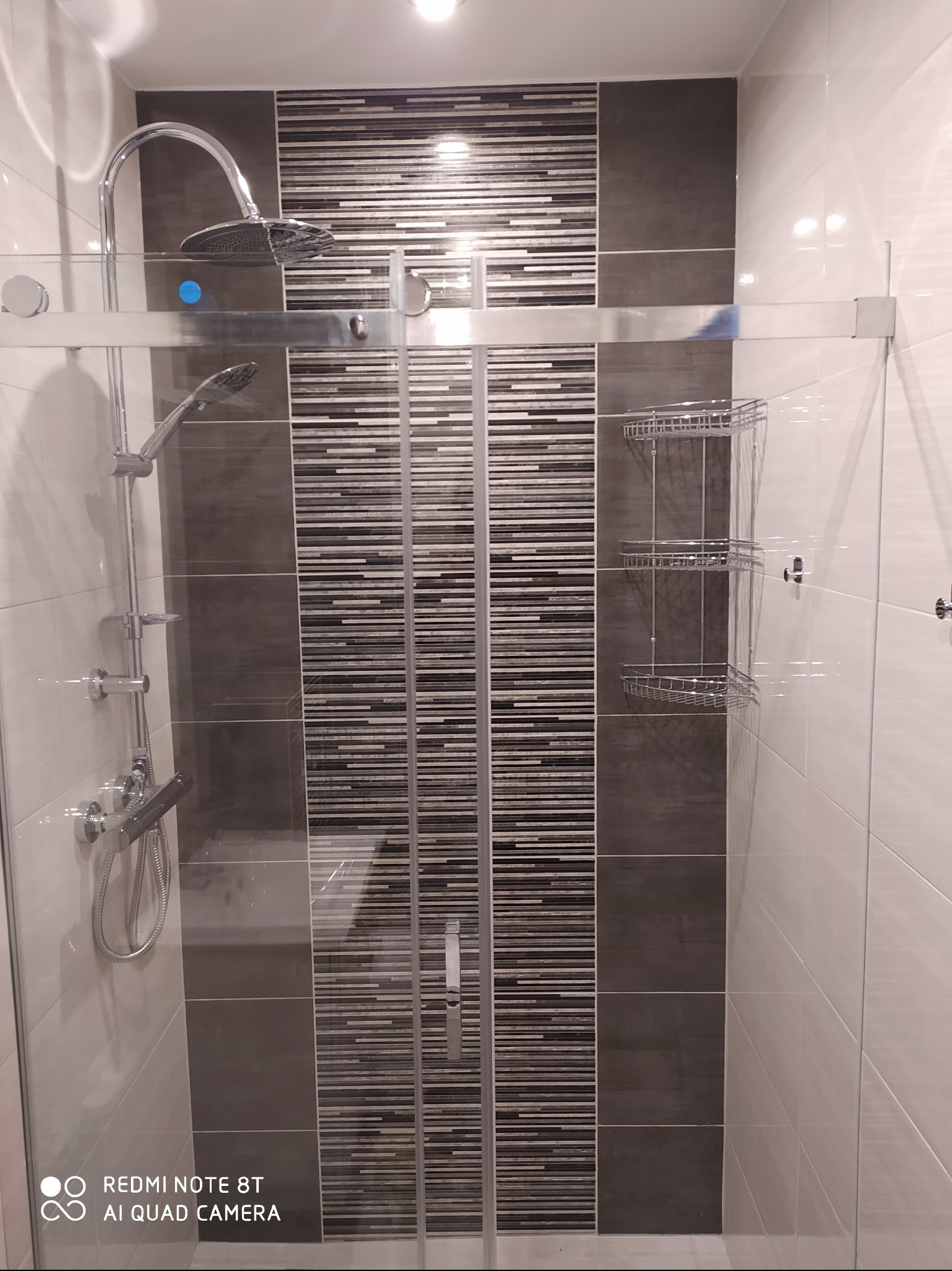 Nowoczesna kabina prysznicowa z chromowaną armaturą, szklanymi drzwiami przesuwnymi i półkami narożnymi na tle płytek w odcieniach szarości i bieli.