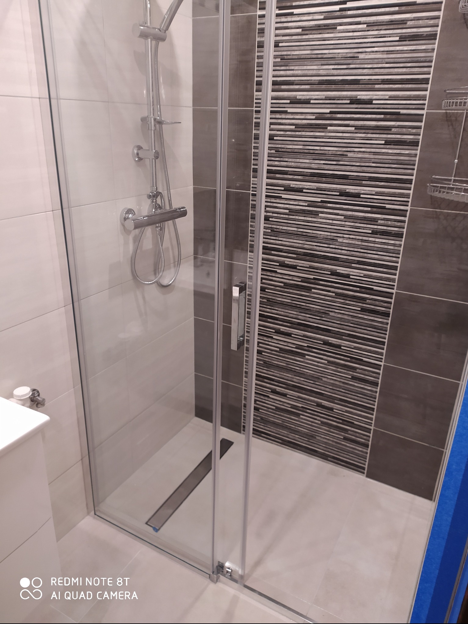 Nowoczesna kabina prysznicowa z przeszklonymi drzwiami, chromowaną armaturą i odpływem liniowym w podłodze, ściana wyłożona płytkami w odcieniach szarości i bieli.