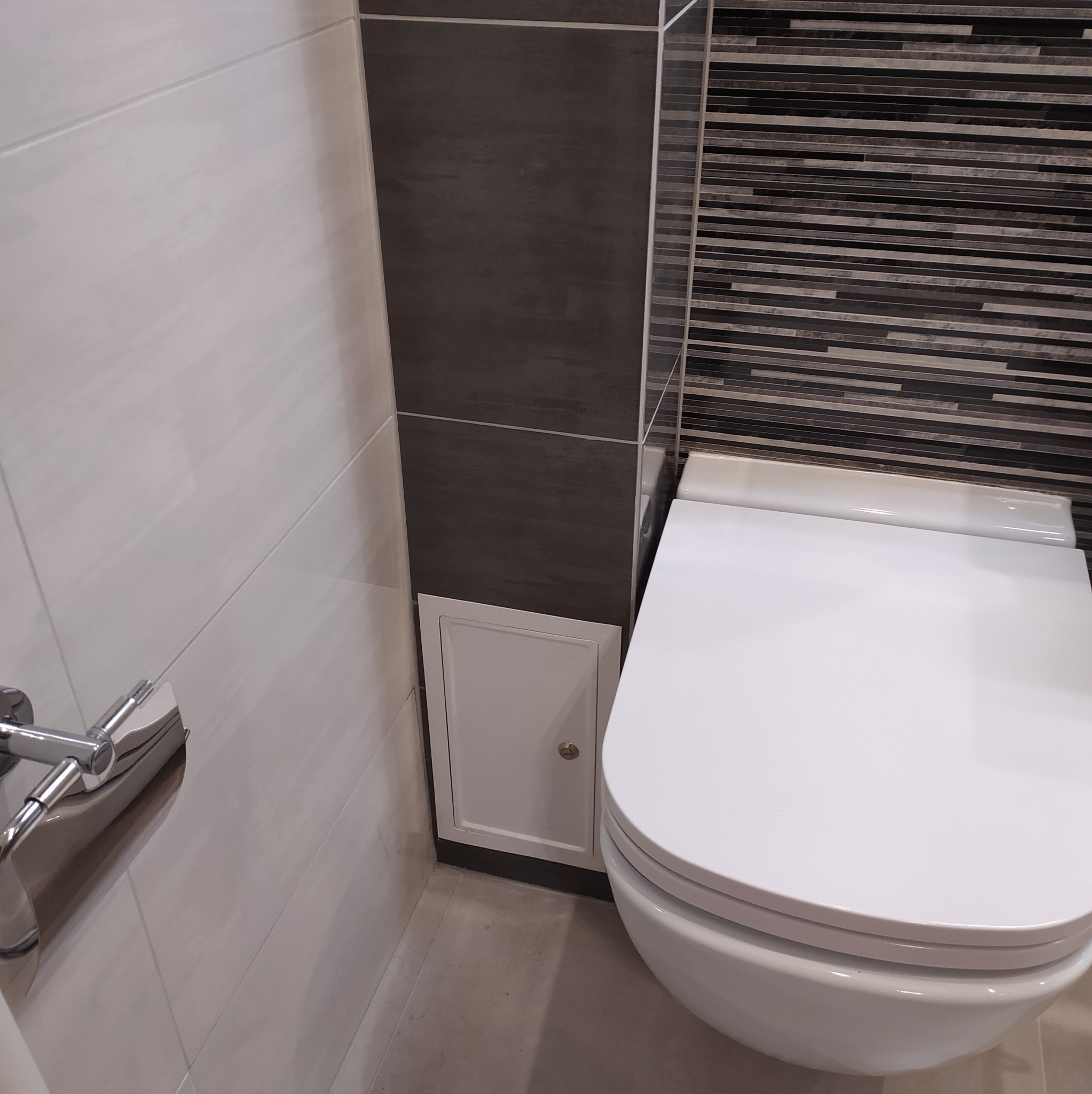 Ściana w łazience z różnymi rodzajami płytek: białe, szare i mozaika, obok podwieszana toaleta i rewizja.