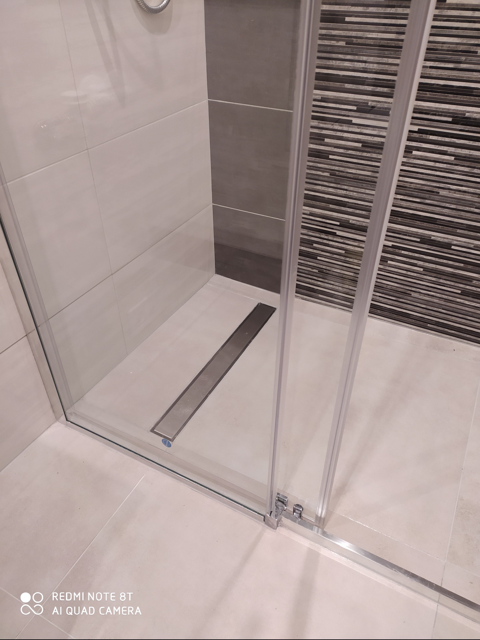 Kabina prysznicowa z odpływem liniowym w nowocześnie wykończonej łazience, widoczne płytki w odcieniach bieli, szarości i czerni.