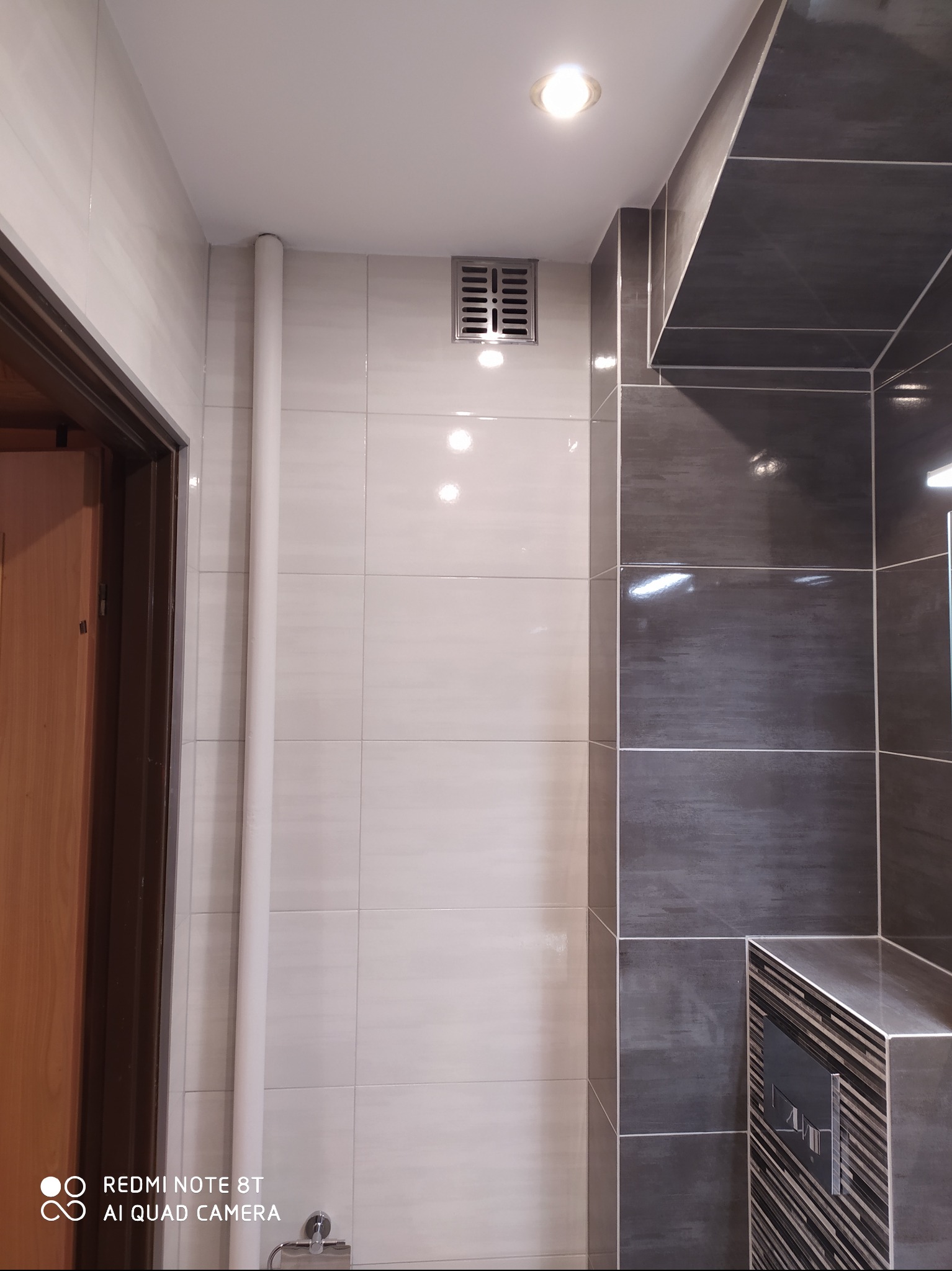 Wykończona łazienka z białymi i szarymi płytkami, widoczna rura pionowa, kratka wentylacyjna i zabudowany stelaż WC.