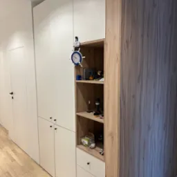 Biało-drewniana zabudowa meblowa z otwartą półką i szufladami, uzupełniona o dekoracyjny zegar w stylu marynistycznym, na tle jasnej podłogi i białych drzwi.