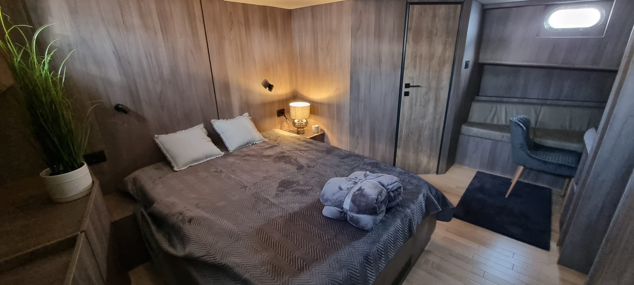 Nowoczesna sypialnia z drewnianymi panelami ściennymi, łóżkiem z szarą pościelą i dekoracjami. Widoczny fragment siedziska z krzesłem w tle.