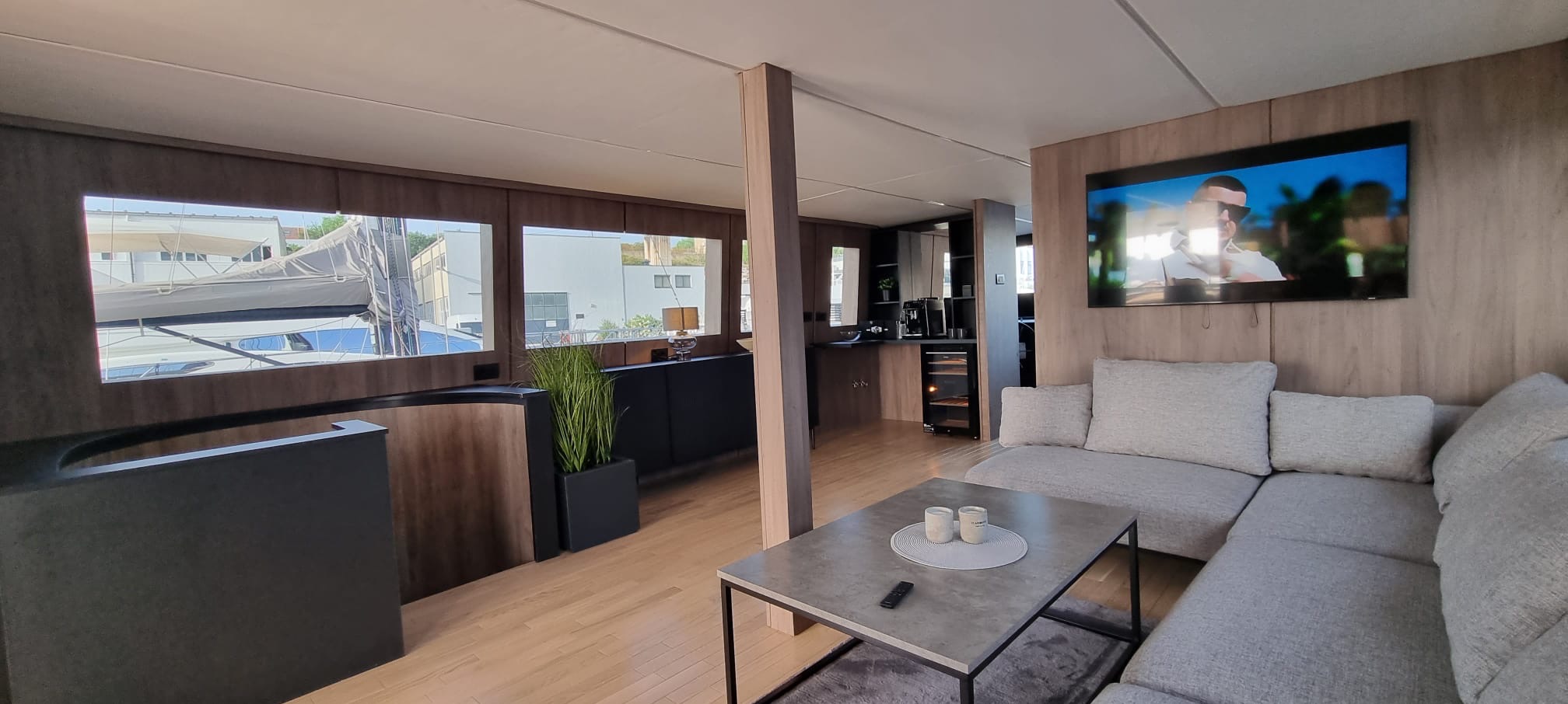 Nowoczesne wnętrze domu na wodzie: salon z szarą sofą narożną, stolikiem kawowym i telewizorem. Widok na jacht przez okna. Drewniane wykończenie ścian i podłóg.