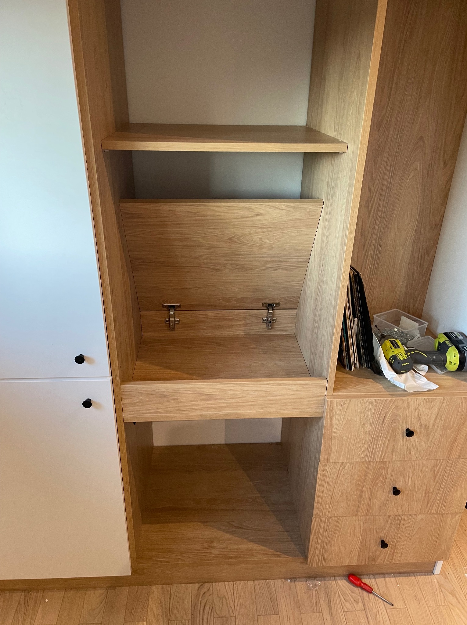 Drewniana szafa z białymi drzwiczkami, widoczne półki i szuflady, narzędzia obok. Detal montażu mebli na wymiar, widoczne zawiasy i elementy konstrukcyjne.