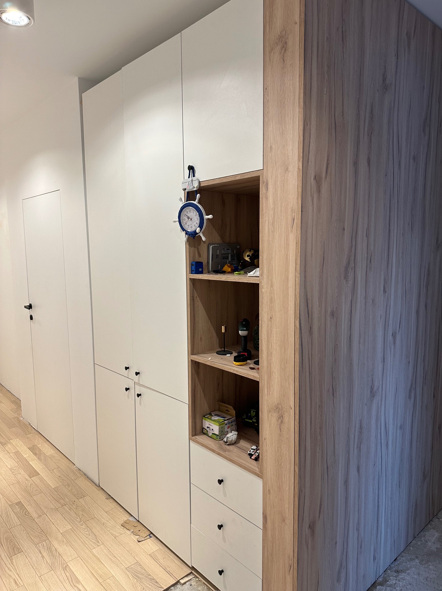 Biało-drewniana zabudowa meblowa z otwartą półką i szufladami, uzupełniona o dekoracyjny zegar w stylu marynistycznym, na tle jasnej podłogi i białych drzwi.