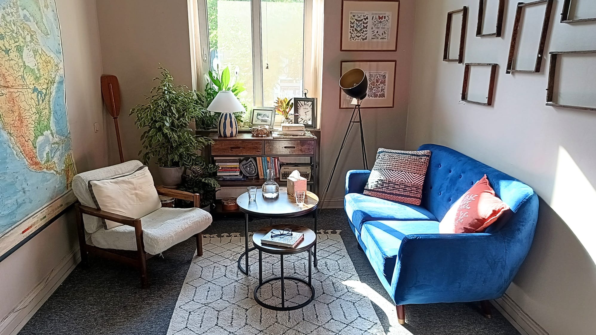 Ustawny pokój z niebieską sofą, stolikiem kawowym z książką i okularami, fotelem, mapą świata na ścianie, lampą stojącą i półką z książkami oraz roślinami.