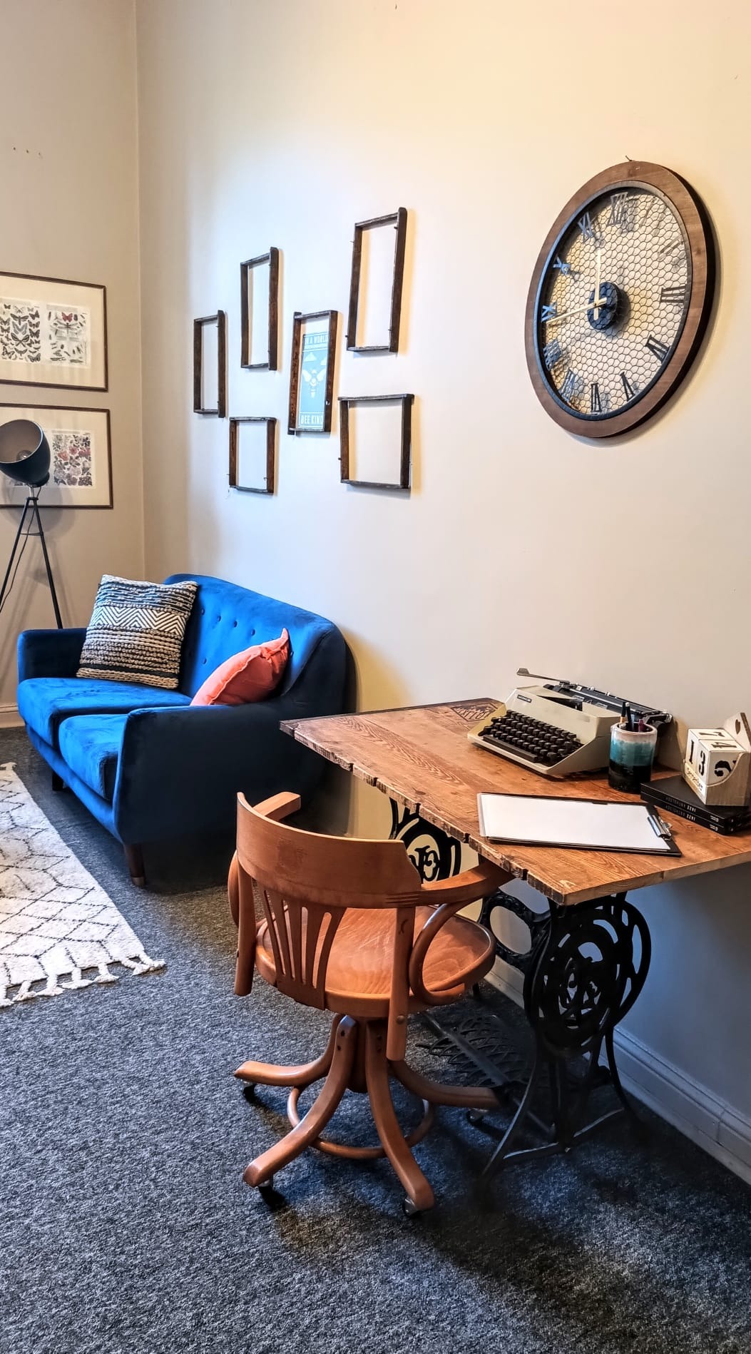 Jasne wnętrze gabinetu z niebieską sofą, drewnianym biurkiem na podstawie maszyny do szycia, starą maszyną do pisania, zegarem ściennym i dekoracjami ścian.