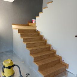 Remonty „Pod klucz” - Montaż drewnianych schodów z widocznym odkurzaczem przemysłowym i puszkami elektrycznymi w ścianie, w trakcie prac wykończeniowych.