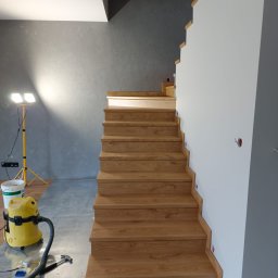 Remonty „Pod klucz” - Drewniane schody z dębowymi stopniami w trakcie montażu, zabezpieczona podłoga, żółty odkurzacz przemysłowy i lampa budowlana w tle, surowa ściana w odcieniach szarości.