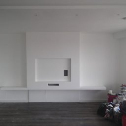 Remonty „Pod klucz” - Białe wnętrze z minimalistycznym kominkiem i półkami, ciemna podłoga, widoczne narzędzia i materiały malarskie w tle, wskazujące na trwające prace wykończeniowe.