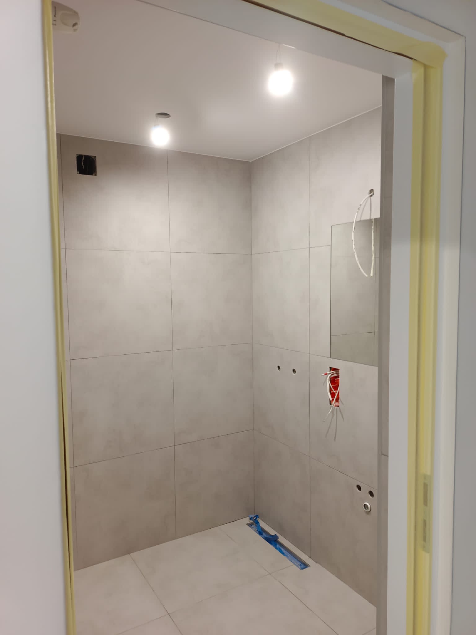 Wnętrze łazienki w trakcie wykańczania, widoczne szare płytki, przygotowane instalacje elektryczne i sanitarne, zabezpieczone folią odpływy liniowe, futryna oklejona taśmą malarską.