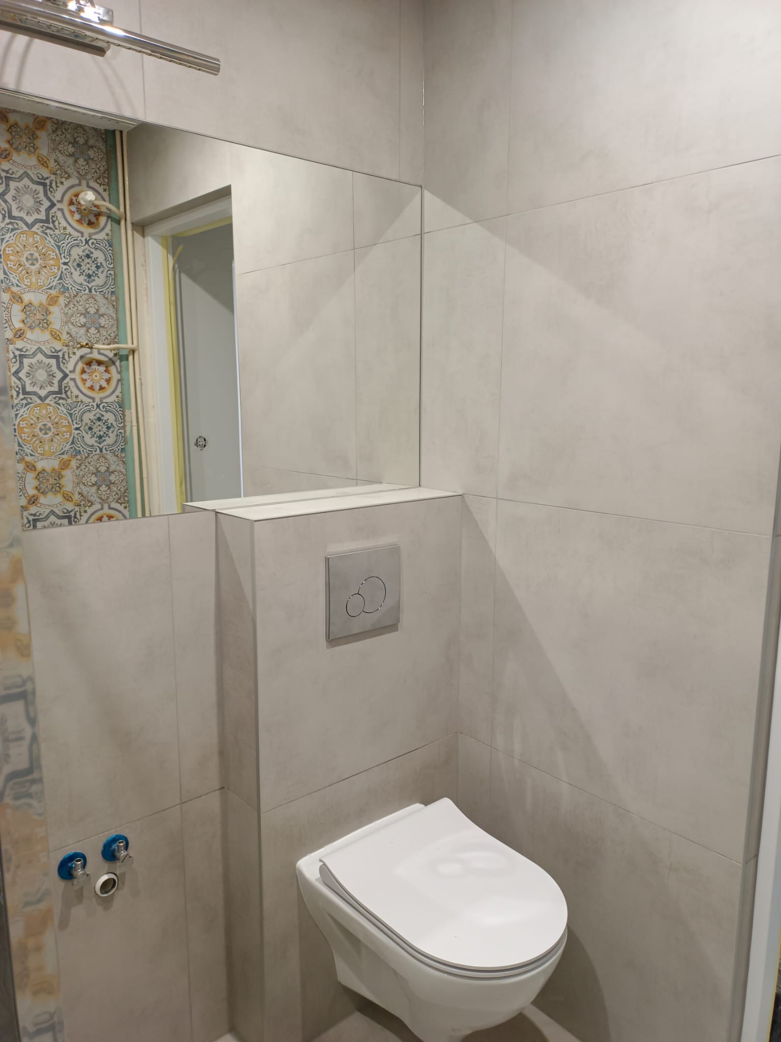 Beżowa łazienka z podtynkową spłuczką, wiszącą toaletą i fragmentem dekoracyjnych płytek widocznych w lustrze.