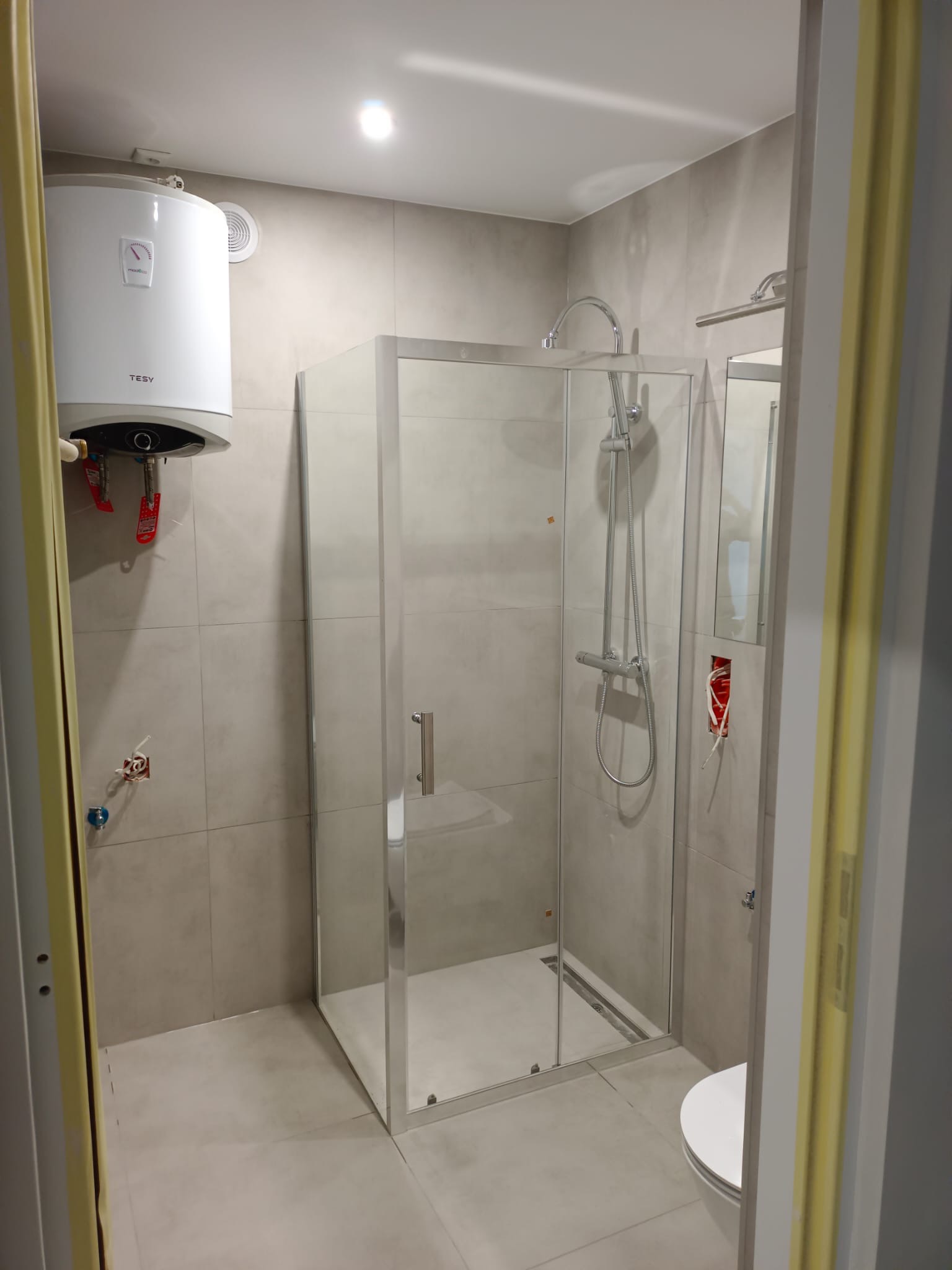 Szara łazienka po remoncie z kabiną prysznicową, bojlerem elektrycznym Tesy na ścianie i widocznymi wystającymi kablami ze ściany w miejscu planowanego gniazdka.