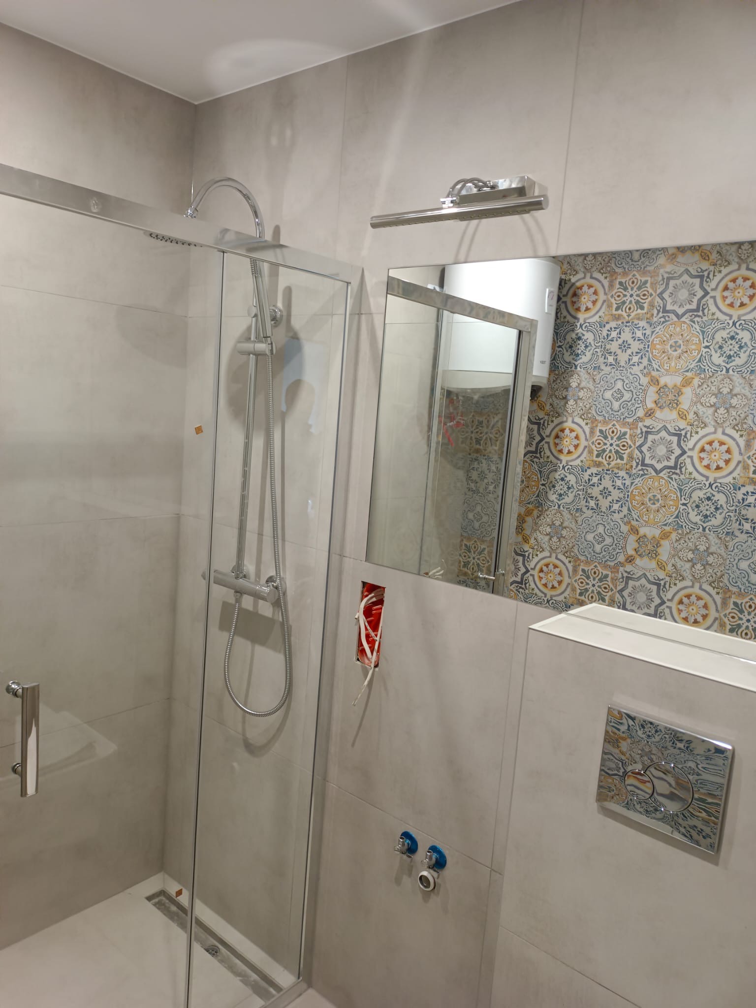 Kabina prysznicowa z chromowaną armaturą i odpływem liniowym, wzorzyste płytki za lustrem, widoczne przyłącza wodne i puszka elektryczna przed wykończeniem.