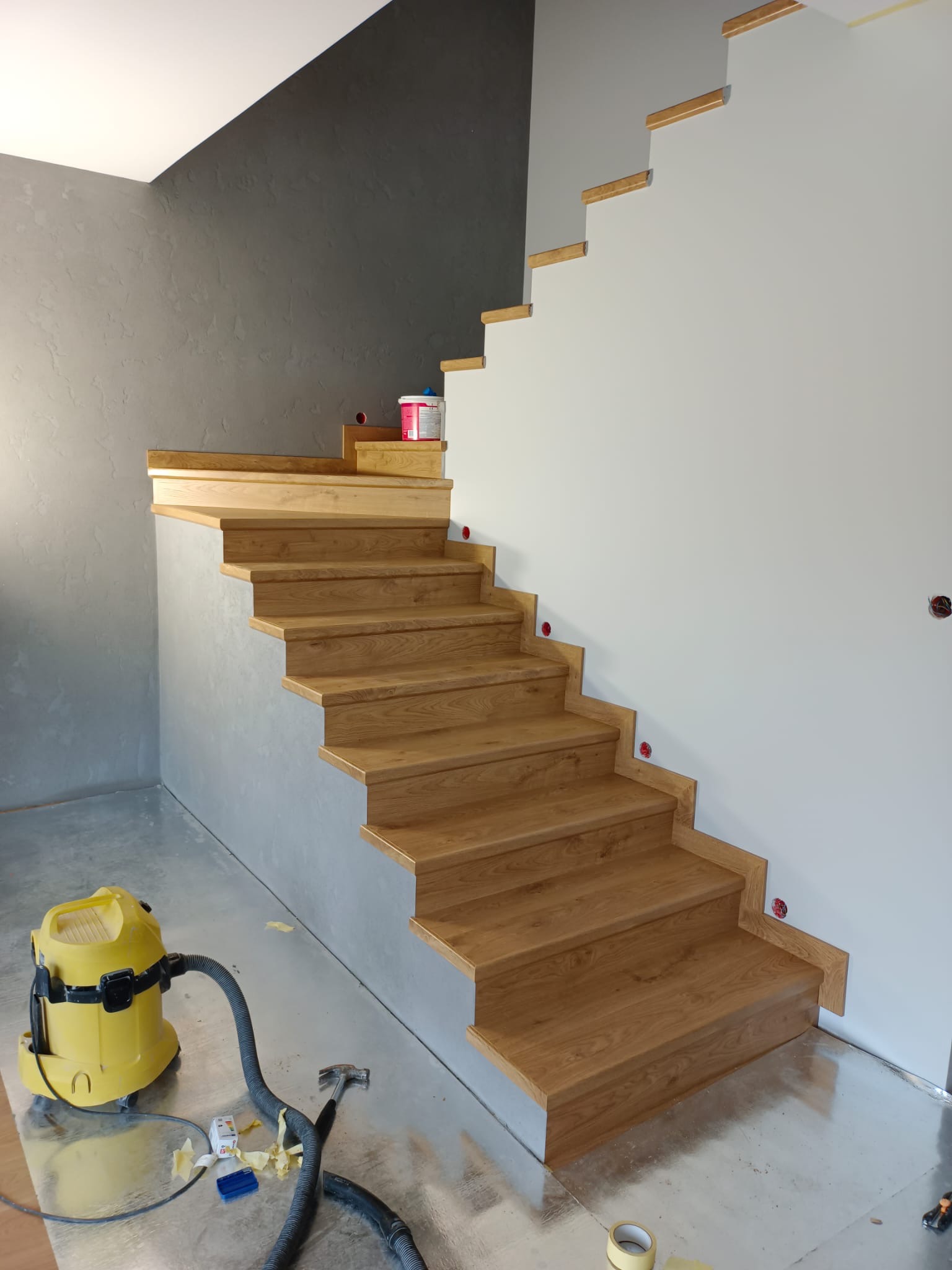 Montaż drewnianych schodów z widocznym odkurzaczem przemysłowym i puszkami elektrycznymi w ścianie, w trakcie prac wykończeniowych.