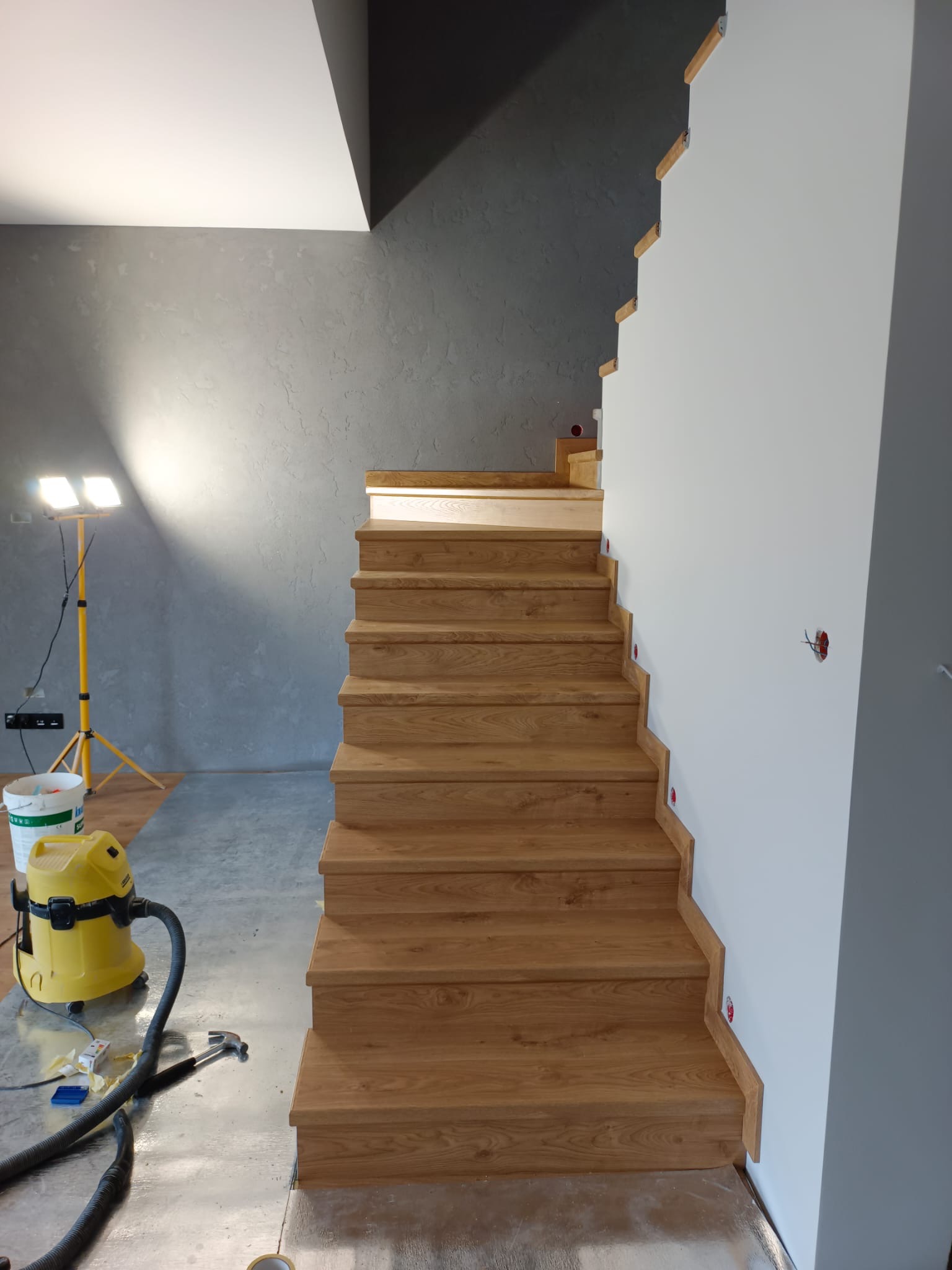 Drewniane schody z dębowymi stopniami w trakcie montażu, zabezpieczona podłoga, żółty odkurzacz przemysłowy i lampa budowlana w tle, surowa ściana w odcieniach szarości.