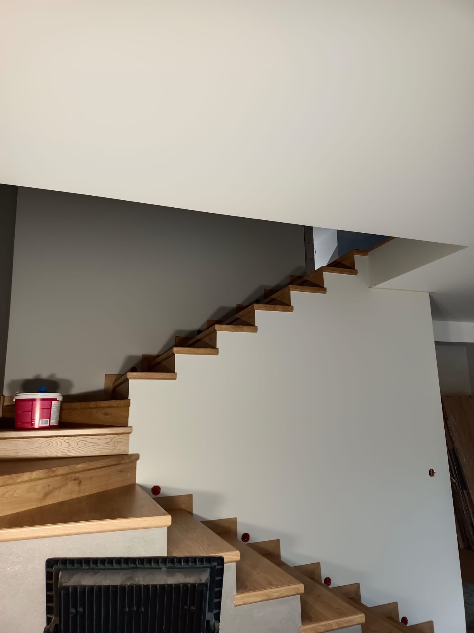 Drewniane schody z pomalowaną na szaro ścianą i puszką farby na jednym ze stopni. Widoczne otwory na oświetlenie w ścianie.