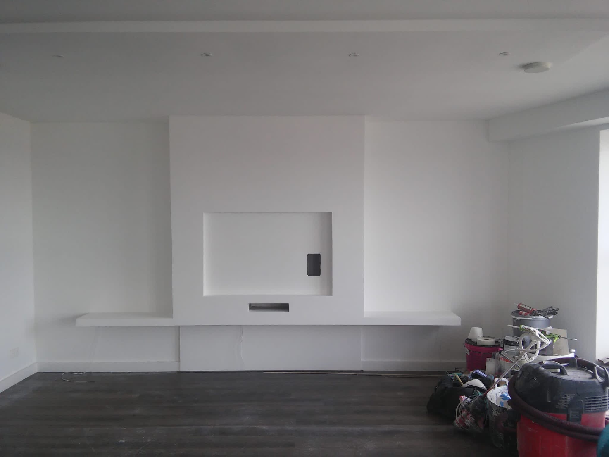 Białe wnętrze z minimalistycznym kominkiem i półkami, ciemna podłoga, widoczne narzędzia i materiały malarskie w tle, wskazujące na trwające prace wykończeniowe.