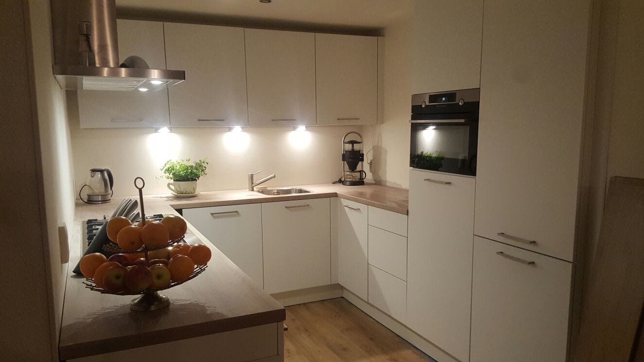 Jasna kuchnia z drewnianym blatem, owocami na paterze i oświetleniem punktowym pod szafkami.