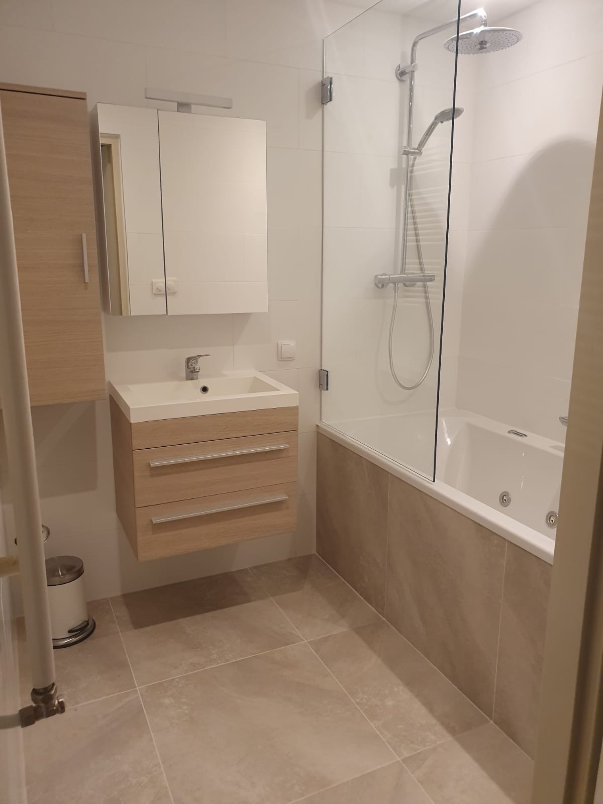 Nowoczesna łazienka z beżowymi płytkami podłogowymi i zabudową w odcieniach drewna, kabina prysznicowa z hydromasażem oddzielona szklaną ścianką.