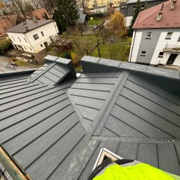 DECKER Paweł Mól - Nowy, grafitowy dach z blachy na tle zabudowy miejskiej, widok z góry na fragment dachu z widoczną strukturą blachy i detalami łączeń, w tle budynki mieszkalne i drzewa.