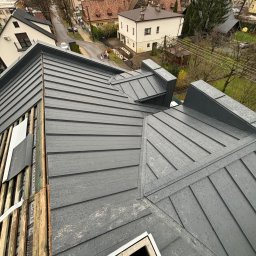 DECKER Paweł Mól - Widok z góry na fragment dachu z blachy na rąbek stojący w kolorze grafitowym, z widocznymi elementami konstrukcji drewnianej i częściowo zamontowaną wentylacją kominową, na tle miejskiej zabudowy...