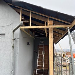 DECKER Paweł Mól - Wznoszenie drewnianej konstrukcji z czarną membraną na dachu domu z szarą elewacją, widoczna drabina, rusztowanie i narzędzia na zewnątrz.