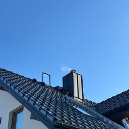 DECKER Paweł Mól - Czarna dachówka ceramiczna na skośnym dachu domu jednorodzinnego, widoczny komin, okno dachowe, rynna i drabina oparta o krawędź dachu na tle błękitnego nieba.