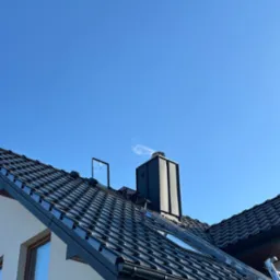 Czarna dachówka ceramiczna na skośnym dachu domu jednorodzinnego, widoczny komin, okno dachowe, rynna i drabina oparta o krawędź dachu na tle błękitnego nieba.