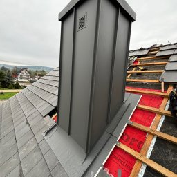 DECKER Paweł Mól - Nowy, szary komin wentylacyjny z blachy na dachu pokrytym szarą dachówką, częściowo zdjętą podczas prac. Widoczne drewniane łaty, membrana dachowa i narzędzia. W tle zabudowa jednorodzinna...