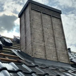 Montaż czarnej dachówki ceramicznej w Limanowej, widoczny komin obłożony imitacją kamienia i przygotowane drewniane łaty pod dach. Częściowo zachmurzone niebo w tle.