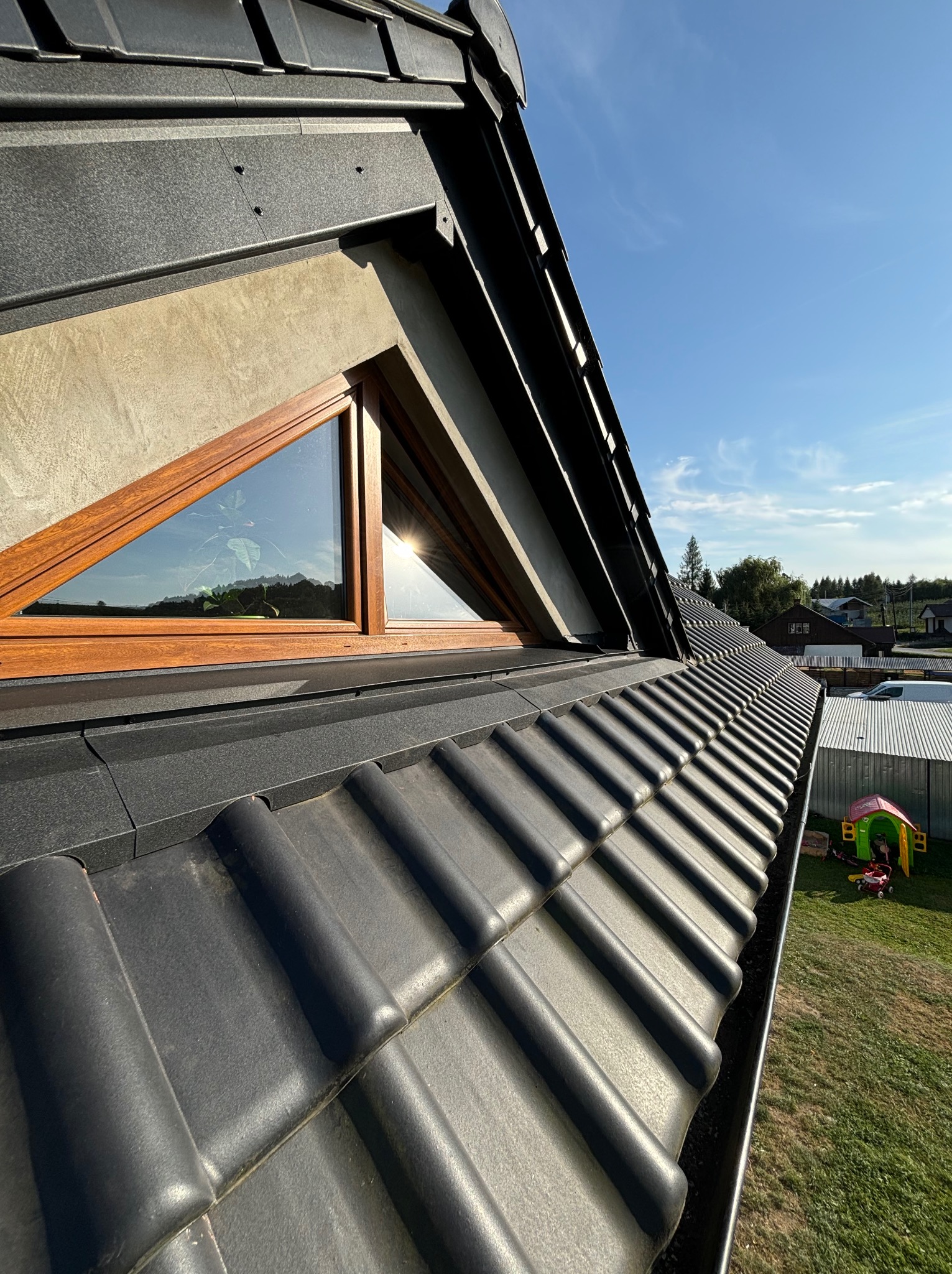 Czarny dach z dachówki ceramicznej z trójkątnym oknem w szczycie, widoczna rynna i otoczenie domu z zielonym trawnikiem w słoneczny dzień.