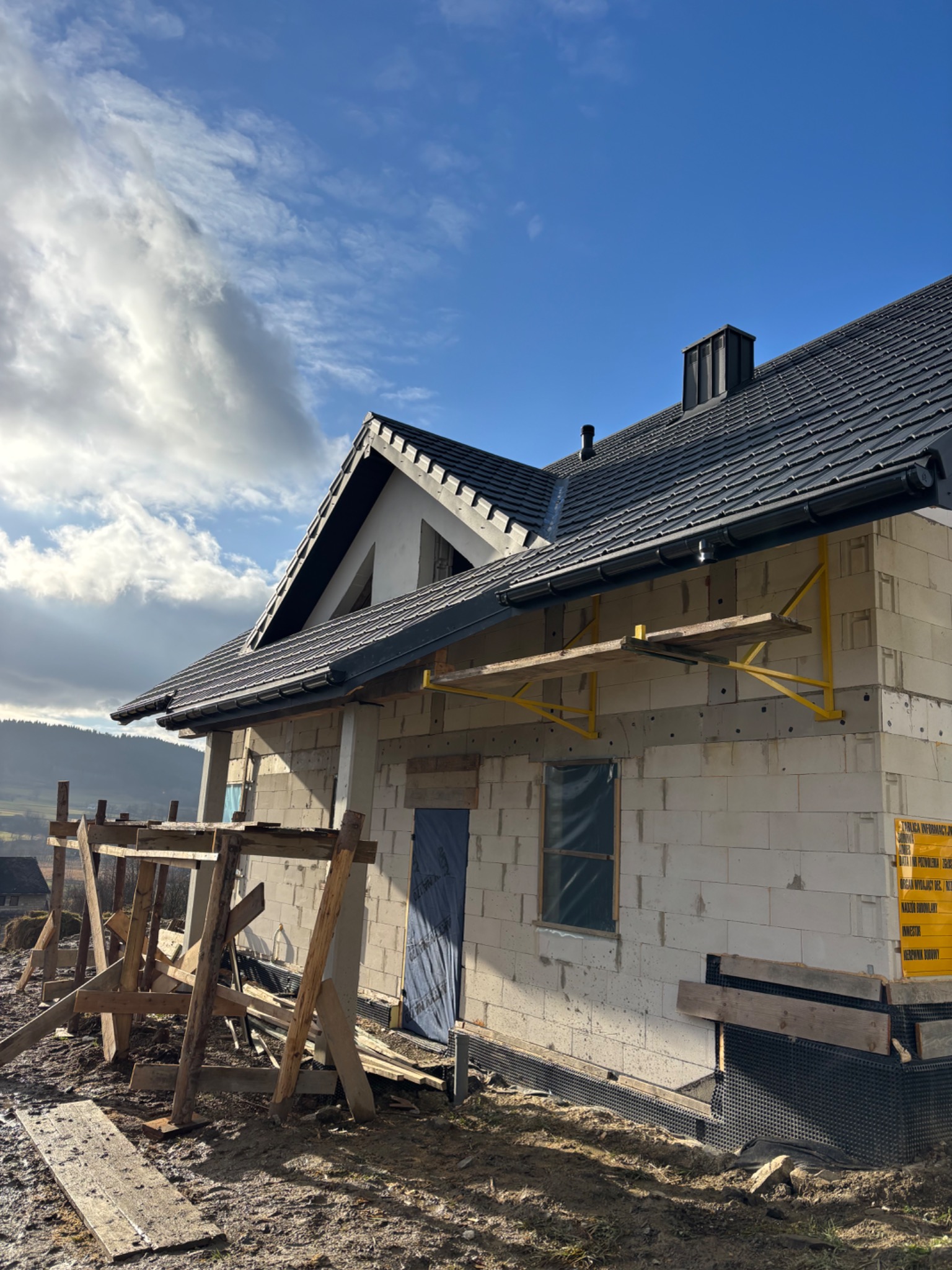 Dom w stanie surowym otwartym z zamontowanym dachem z czarnej dachówki, widoczne rusztowania i folia fundamentowa, w tle pagórkowaty krajobraz pod błękitnym niebem z chmurami.