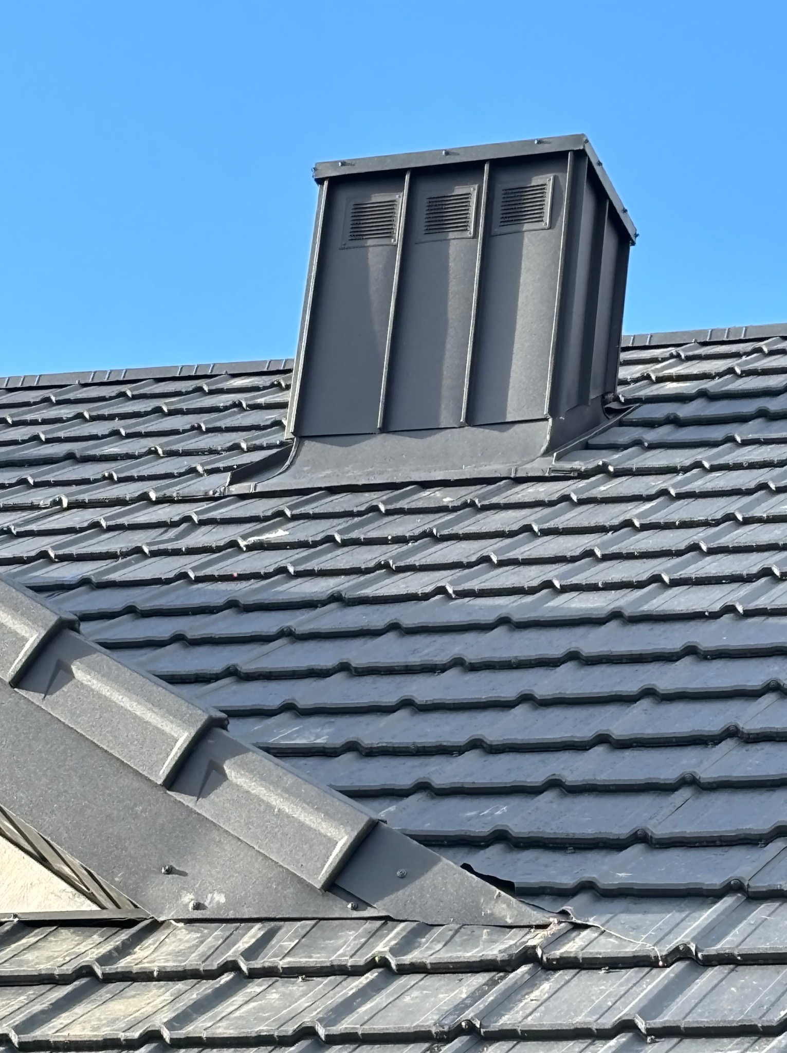 Szary dach z dachówki ceramicznej z obróbką blacharską komina z trzema otworami wentylacyjnymi na tle błękitnego nieba.