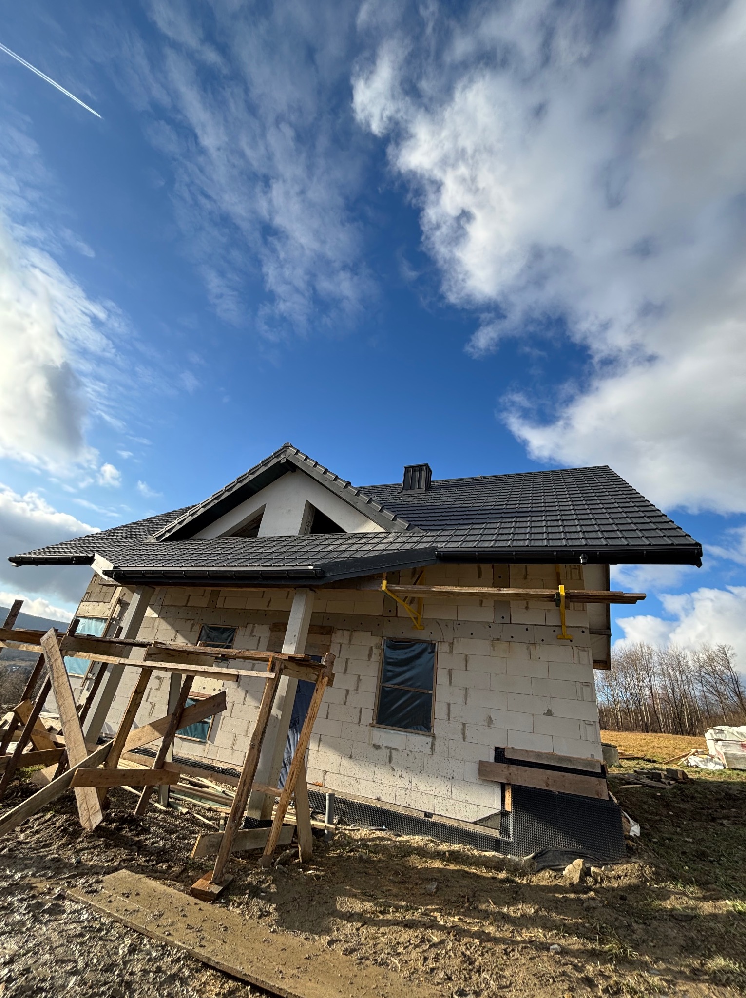 Dom jednorodzinny w trakcie budowy, z widoczną konstrukcją dachu pokrytą czarną dachówką, zabezpieczone okna folią, rusztowanie drewniane w tle błękitne niebo z chmurami i smugą kondensacyjną...