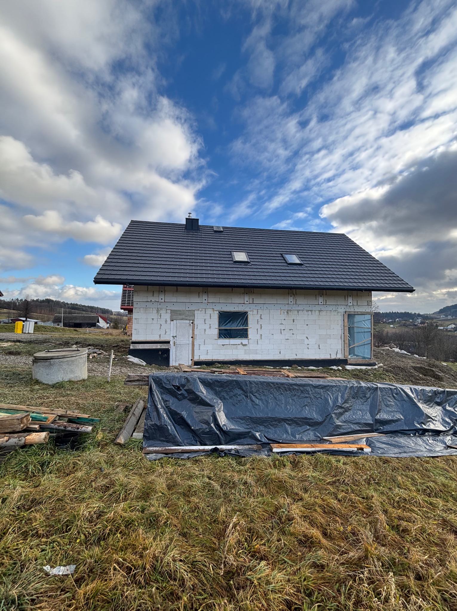 Surowy stan budynku z szarym dachem z dachówki ceramicznej i oknami dachowymi, widok na budowę domu z pustaków, w tle krajobraz wiejski pod pochmurnym niebem.