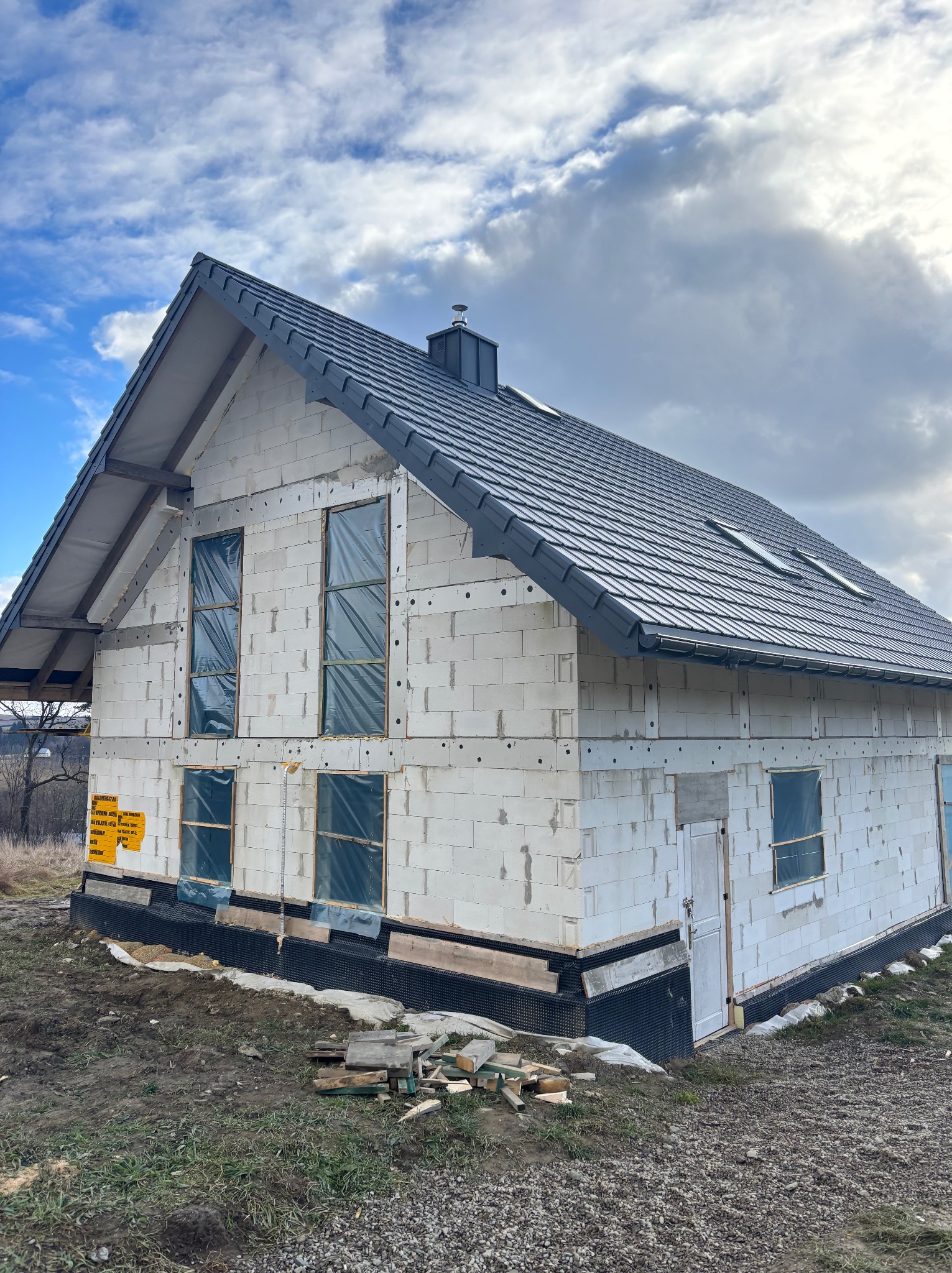 Stan surowy otwarty domu jednorodzinnego w Limanowej z widoczną konstrukcją dachu pokrytą szarą dachówką, okna zabezpieczone folią, ściany z białego bloczka i przygotowany teren wokół budynku.
