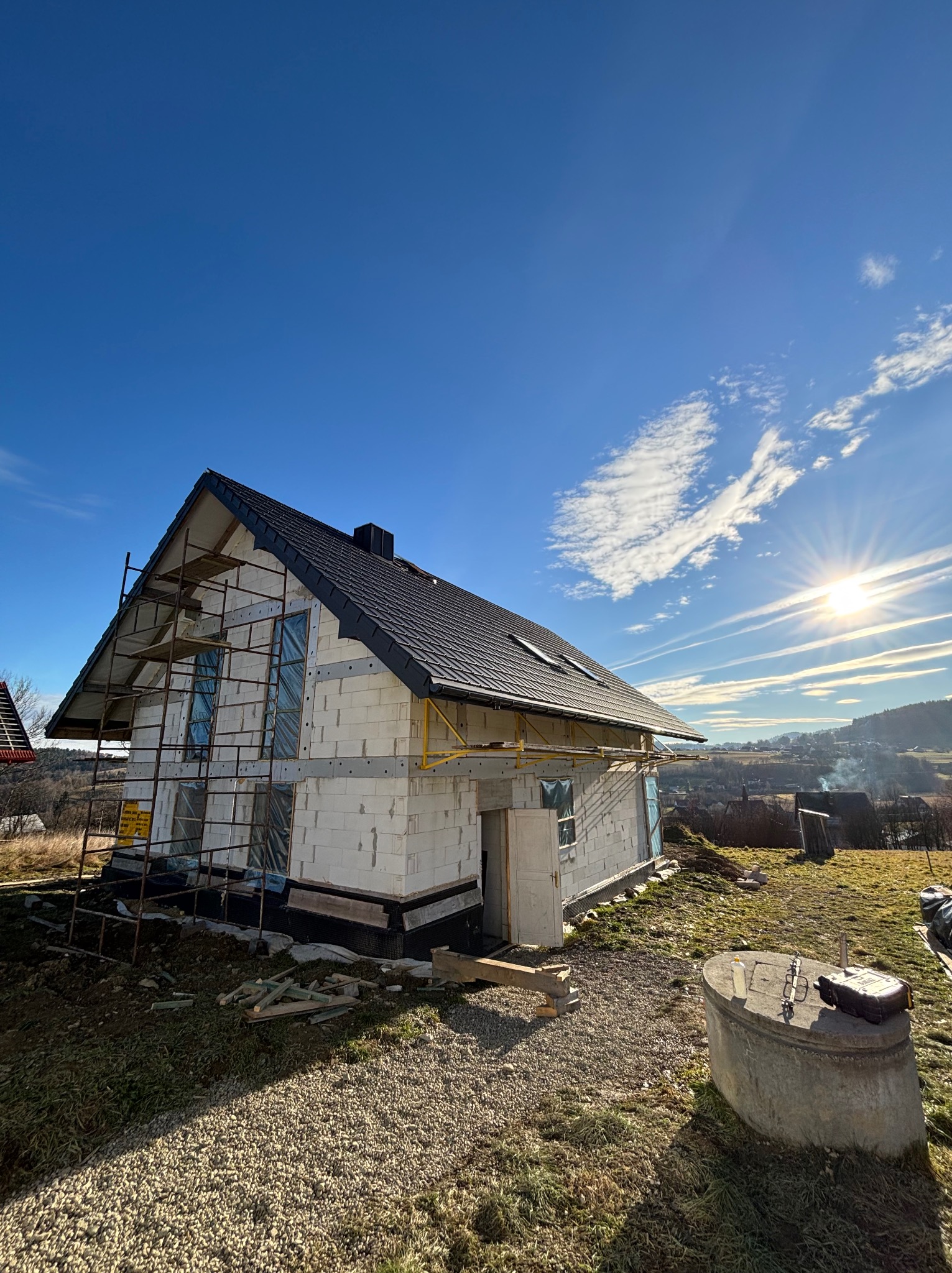 Budowa domu jednorodzinnego w stanie surowym zamkniętym z czarną dachówką, widoczne rusztowania i okna zabezpieczone folią, na tle błękitnego nieba z chmurami i słońcem.