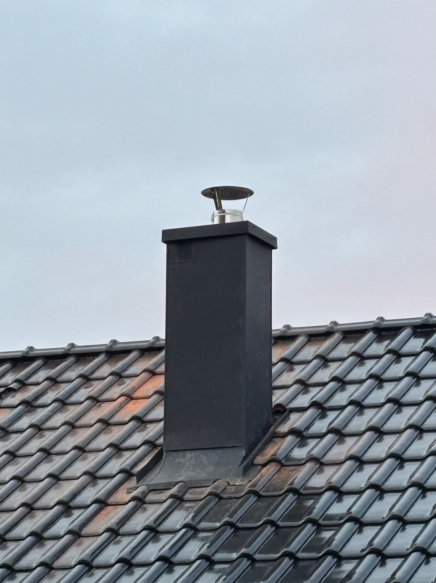Czarny, metalowy komin z nasadą na dachu pokrytym ciemną dachówką ceramiczną, widoczne ślady wilgoci po deszczu.