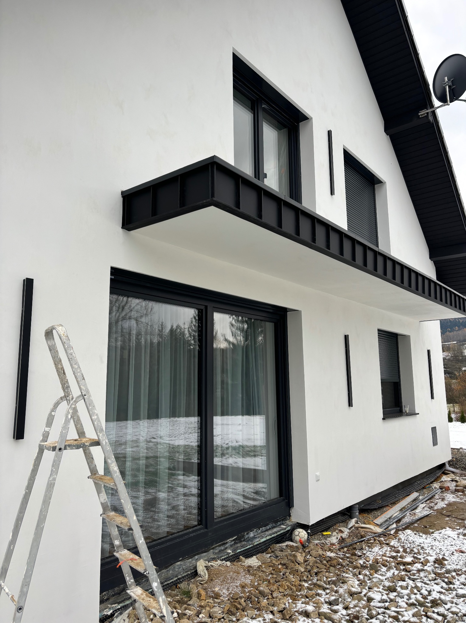 Nowoczesny dom z białymi ścianami i czarnymi detalami, widoczna drabina oparta o ścianę, teren wokół budynku w trakcie prac wykończeniowych.
