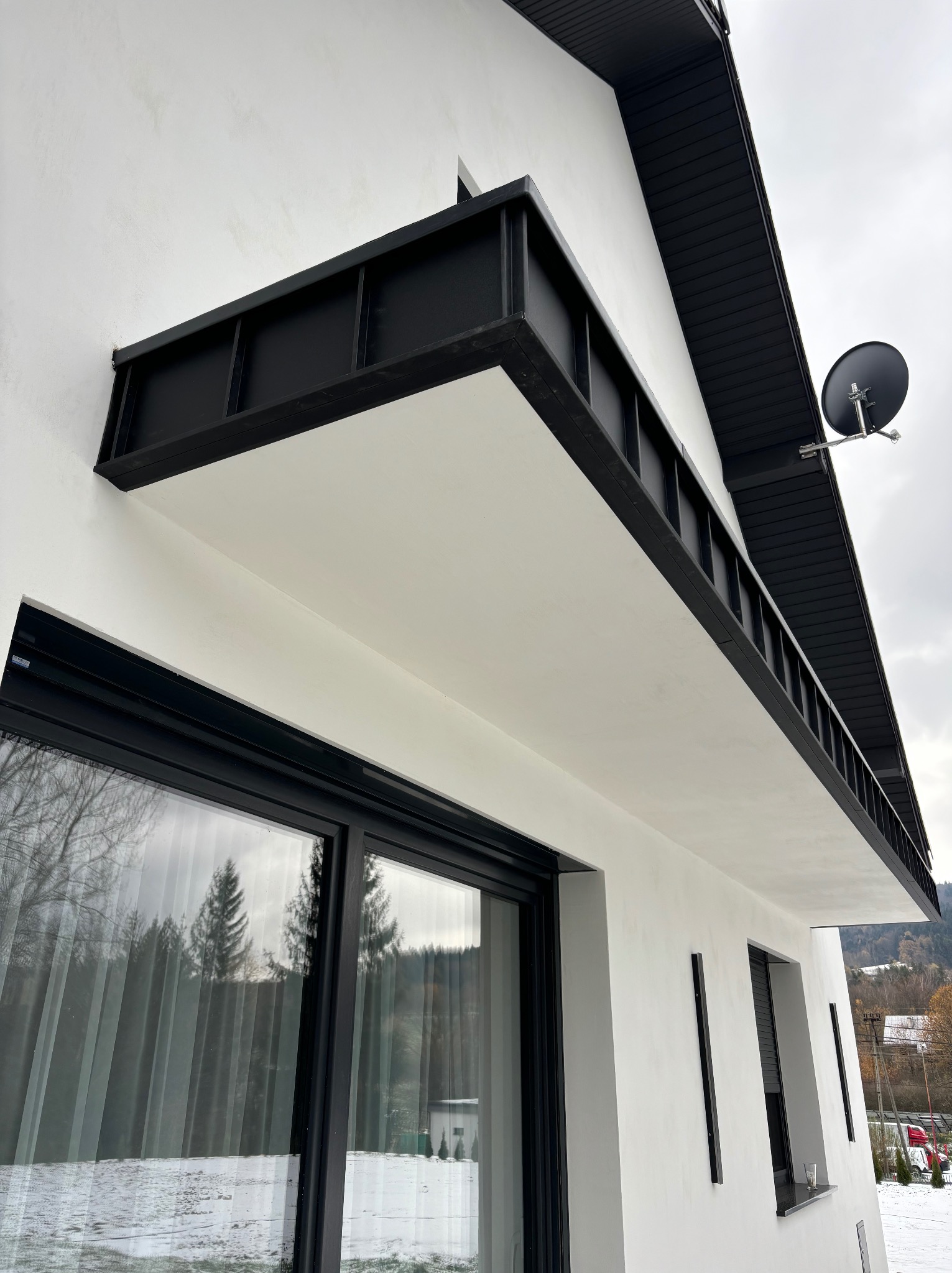 Nowoczesny dom z białymi ścianami i czarnym dachem z blachy, widoczny balkon z czarną balustradą i antena satelitarna na elewacji, za oknem widok na zimowy krajobraz z drzewami.