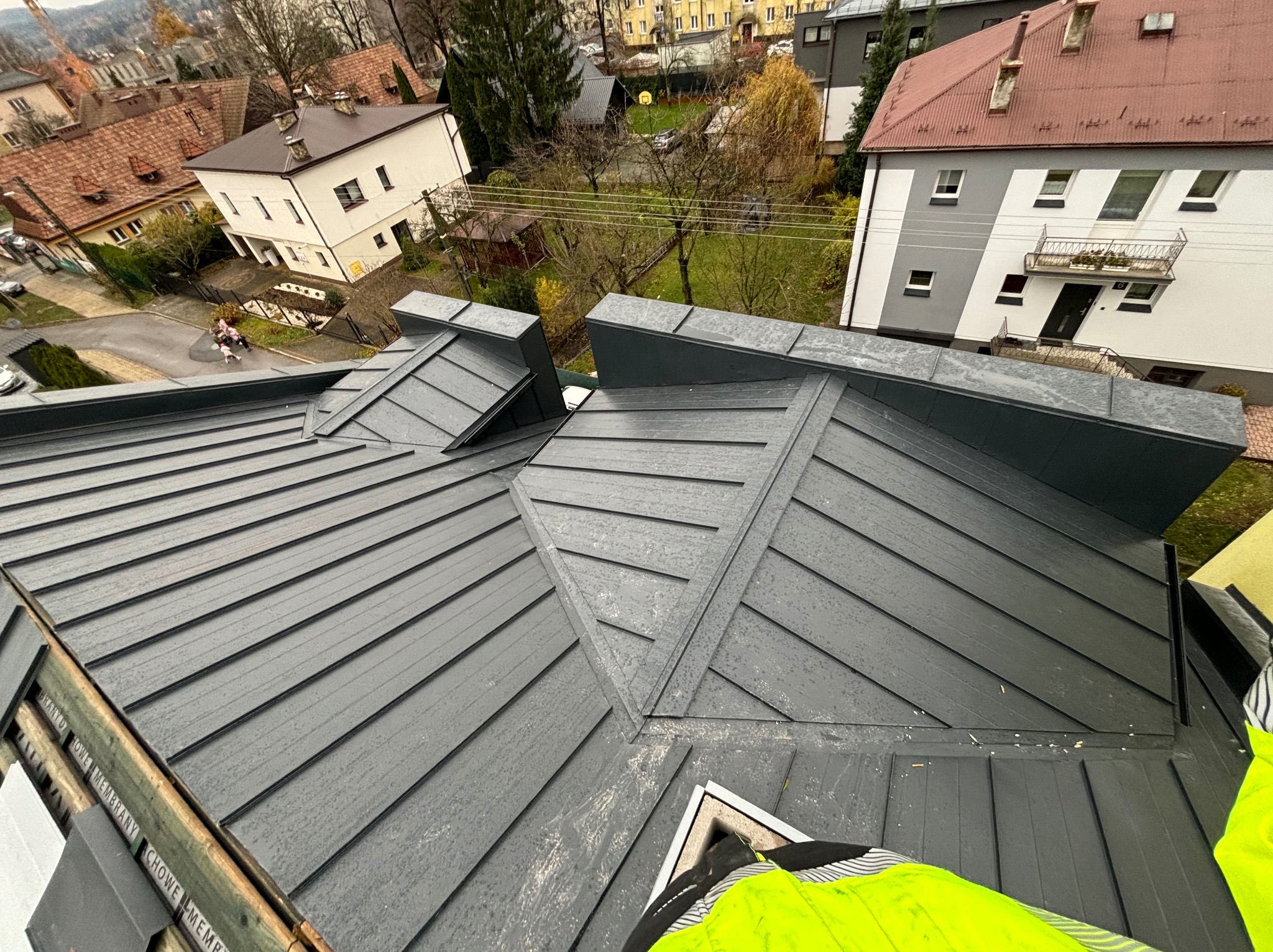 Nowy, grafitowy dach z blachy na tle zabudowy miejskiej, widok z góry na fragment dachu z widoczną strukturą blachy i detalami łączeń, w tle budynki mieszkalne i drzewa.