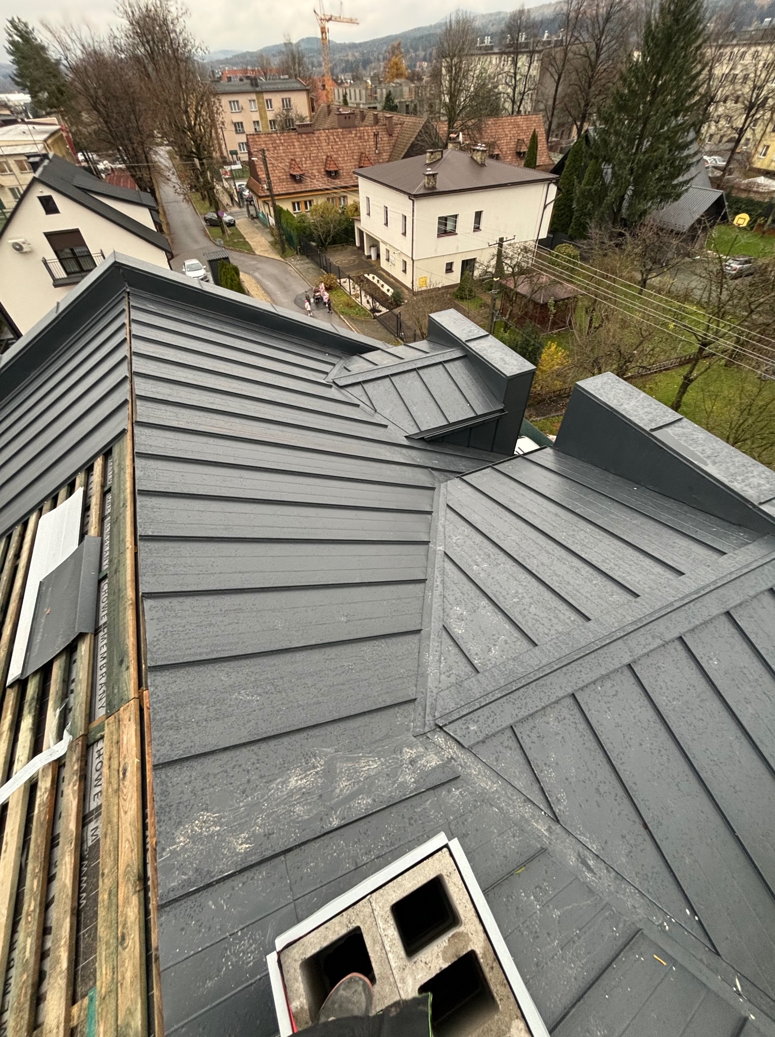 Widok z góry na fragment dachu z blachy na rąbek stojący w kolorze grafitowym, z widocznymi elementami konstrukcji drewnianej i częściowo zamontowaną wentylacją kominową, na tle miejskiej zabudowy...