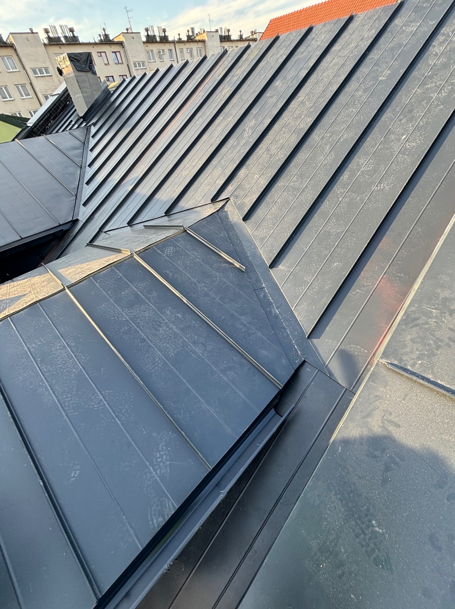 Szare blaszane pokrycie dachowe na budynku wielorodzinnym, widoczne z góry pod kątem, z elementami obróbek blacharskich i kominem w tle.