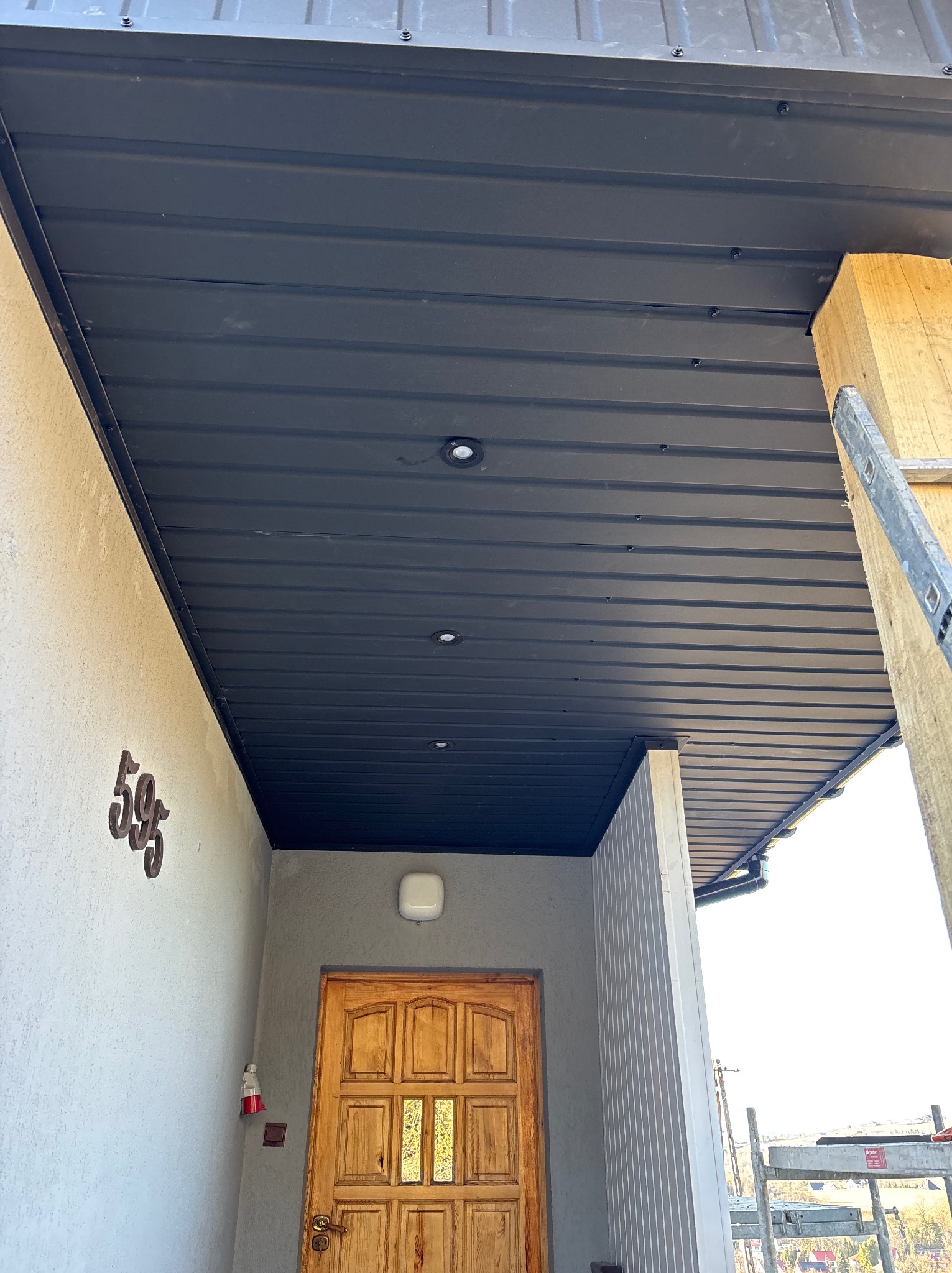 Ciemna, metalowa podbitka dachowa z wbudowanymi punktami świetlnymi, widoczna z dołu, z fragmentem drabiny opartą o ścianę i numerem domu '595' na elewacji.