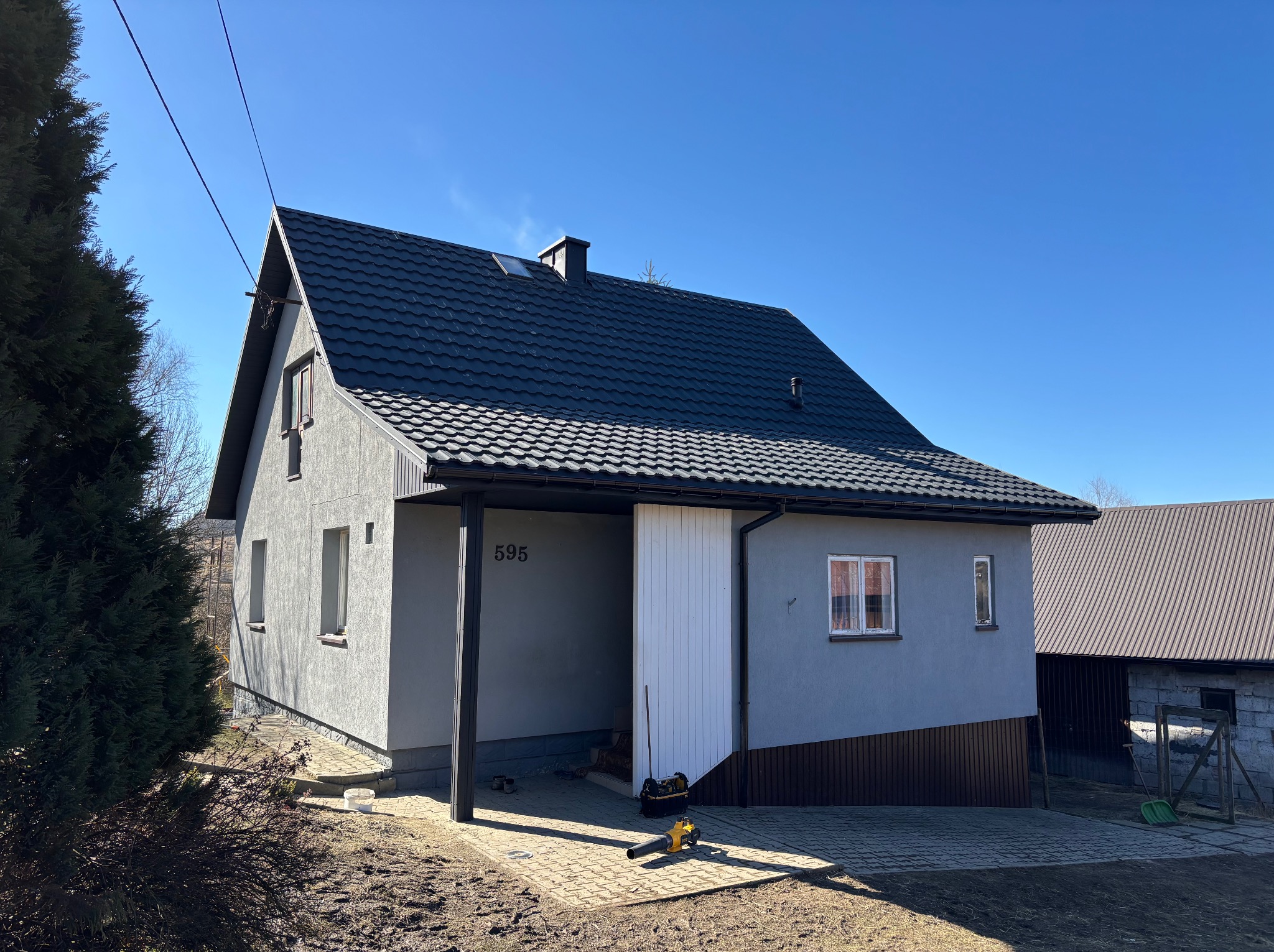 Nowoczesny dom jednorodzinny z ciemnoszarą dachówką, numerem 595 nad wjazdem i dmuchawą do liści na kostce brukowej przed wjazdem.