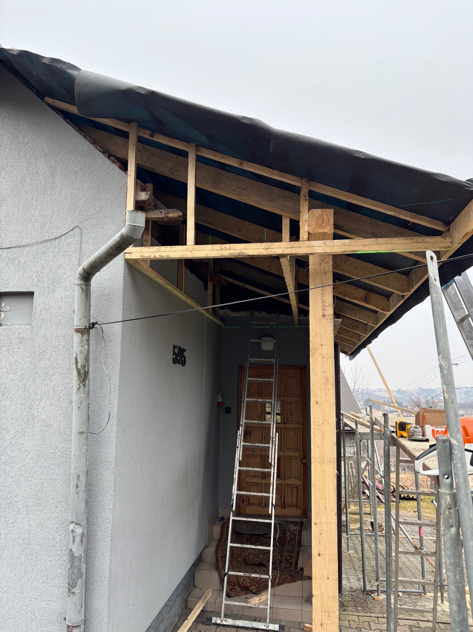 Wznoszenie drewnianej konstrukcji z czarną membraną na dachu domu z szarą elewacją, widoczna drabina, rusztowanie i narzędzia na zewnątrz.